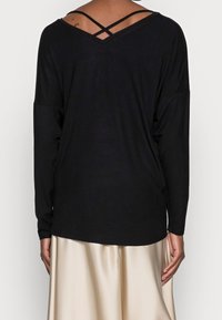 Schwarzes langärmliges Oberteil mit V-Ausschnitt und gekreuztem Trägerdetail am Rücken, ausgestattet mit einer lockeren Passform und weichem Stoff.