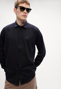 Svart långärmad skjorta med ett subtilt mönster, krage och 'BOSS'-logotyp på bröstet, kombinerad med mörka solglasögon.