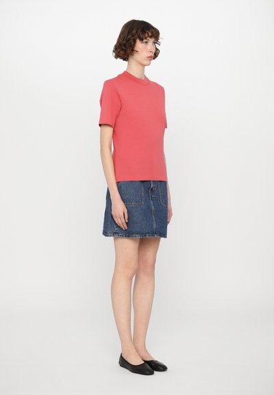 Coral short-sleeve top, relaxed fit, paired with a knee-length denim skirt. Model wearing black flats, standing against a plain white background.