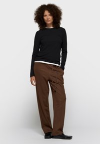 Top negro de manga larga sobre una camisa blanca, combinado con pantalones marrones de corte holgado con cordón y logo. Calzado negro.