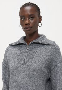 Maglione grigio in lana con colletto a zip quarti, texture a coste e un taglio ampio e confortevole. Dettagli delle cuciture visibili sul davanti.