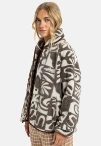 Veste en polaire marron et crème, avec des motifs abstraits. Texture douce, bordures marron et boutons-pression sur le devant. Comprend des poches latérales.
