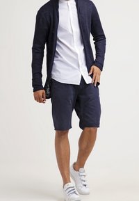 Námořnický pletený cardigan, bílá košile na knoflíky, námořnické šortky a bílé tenisky. Cardigan má otevřený přední díl, volný střih a strukturovanou tkaninu.