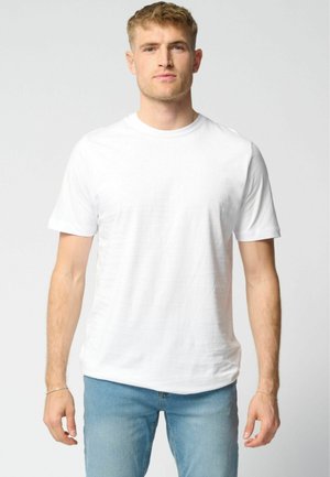 Fiatal férfi rövid világosbarna hajjal, sima fehér rövid ujjú pólóban és világoskék farmerral, semleges háttér előtt áll.