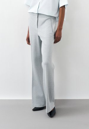 Person som bærer lysegrå harlekinmønstrede vidbukser, hvit innsatt bluse og svarte sko med spiss tupp, står mot en hvit vegg.