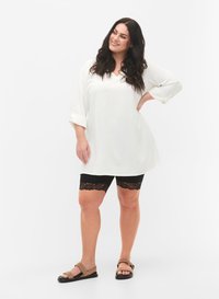 Pitkähiuksinen nainen, jolla on valkoinen pitkähihainen tunika, mustat pitsireunaiset pyöräilyshortsit ja vaaleat sandaalit, hymyillen ja käsi lanteilla.