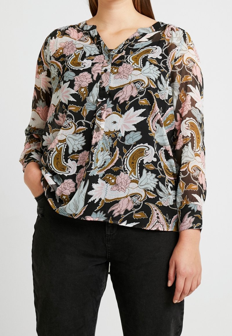 Femme portant une blouse noire à manches longues avec des motifs floraux rose, bleu et marron, associée à un pantalon noir et une main dans la poche.
