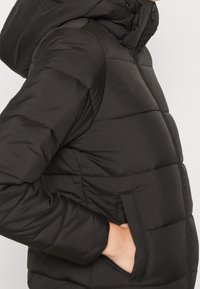 Doudoune noire avec capuche, présentant un design matelassé, une texture lisse et des poches latérales. Fermeture visible au niveau du cou.