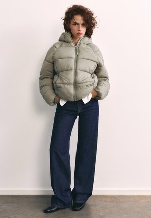 Femme aux cheveux bouclés portant une veste matelassée grise, un jean large foncé, des chaussures noires et une chemise blanche, debout contre un mur blanc uni.