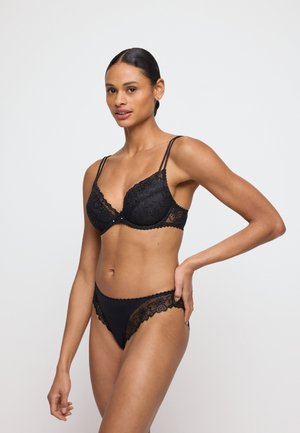 Nainen seisoo käsi lanteilla, musta pitsiliivit ja niihin sopivat alushousut päällä yksiväristä valkoista taustaa vasten.