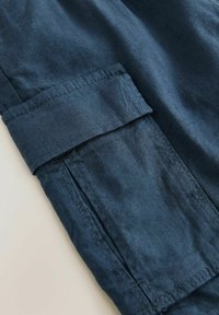 Tessuto blu scuro con una superficie liscia, caratterizzato da polsini arrotolati e dettagli di cucitura visibili. Il design include tasche laterali per la funzionalità.