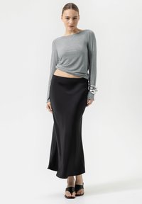 Haut gris à manches longues avec une coupe ample, associé à une jupe maxi en satin noir. Le mannequin porte des sandales noires à enfiler et des bracelets en argent.