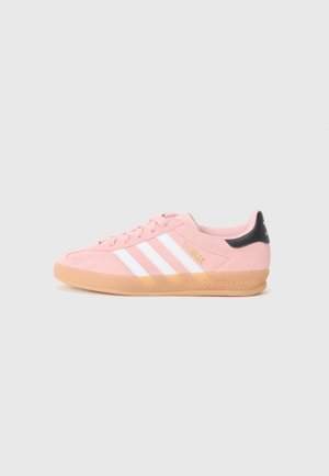 Różowy sneaker Adidas Gazelle z białymi paskami, gumową podeszwą i czarnym elementem na pięcie, pokazany z boku na jednolitym tle.
