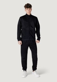 Tuta sportiva nera realizzata in tessuto leggero. La giacca presenta un colletto alto e una chiusura con zip; i pantaloni hanno dettagli a pannello e polsini elasticizzati.