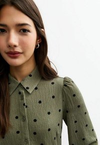Blouse verte texturée avec des pois noirs, devant boutonné et un col. Les manches bouffantes ajoutent du volume au design.