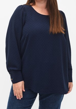 Femme portant un chemisier texturé bleu marine à manches longues et un jean bleu, debout avec une main visible sur le côté.