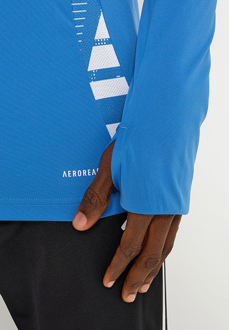 Mano descansando cerca de la cadera, vistiendo una camiseta deportiva de manga larga azul con agujero para el pulgar y pantalones negros con una franja blanca.