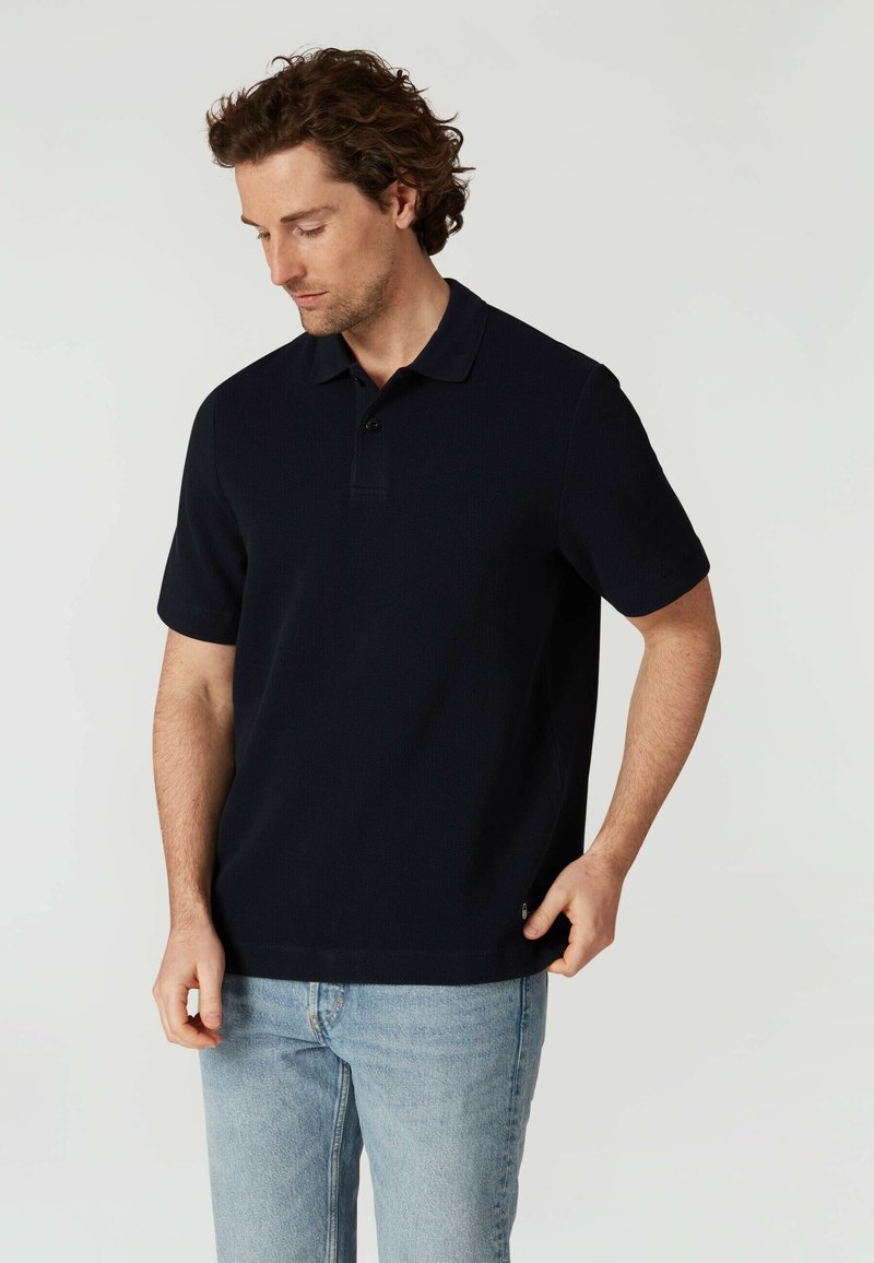 Mörkblå polo med krage, korta ärmar och treknappsstängning; tillverkad i strukturerad tyg; kombinerad med ljusblå jeans.