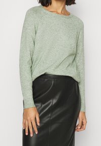 Ljusgrön, texturerad stickad tröja med rund halsringning och ribbade ärmslut, kombinerad med en svart läderskjort som har en slät yta.