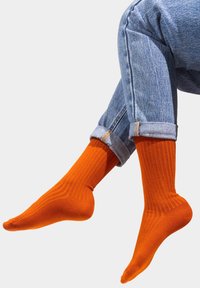 Pomarańczowe skarpetki w żebrowany wzór, wykonane z miękkiego materiału, o długości do połowy łydki, noszone z podwiniętymi jasnoniebieskimi dżinsami.