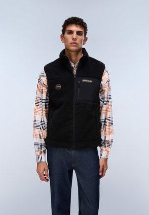 Young man wearing a black zip-up fleece vest over a plaid shirt and dark jeans standing against a plain light background.