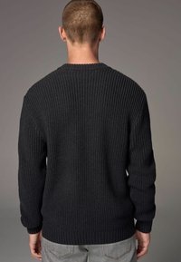 Une personne se tient de dos, portant un pull en tricot sombre avec un motif texturé, complété par un jean gris clair, le tout sur un fond neutre.