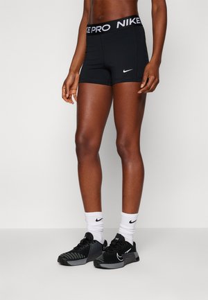 Athlète portant un short Nike Pro noir, des chaussettes blanches Nike et des chaussures d'entraînement Nike noires sur un fond uni.