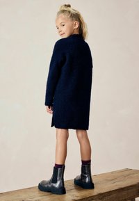 Manteau en tricot moelleux bleu marine avec un col haut, de conception mi-longue, porté sur des chaussettes beiges avec des bottines noires à semelles chunky.