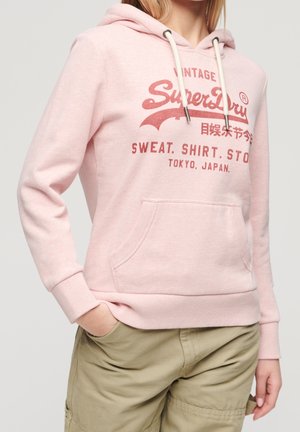 Kobieta ubrana w jasnoróżową bluzę z kapturem z czerwonym logo "SuperDry" i beżowe spodnie, ręka w kieszeni, na jednolitym jasnym tle.