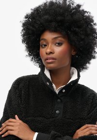 Black fuzzy fleece pullover with a high collar and snap buttons. Features a soft texture, contrasting fabric, and a relaxed fit.