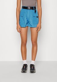 Shorts légers bleus avec deux poches avant, une ceinture noire ajustable et une texture lisse. Portés avec des chaussures de sport noires et des chaussettes blanches.