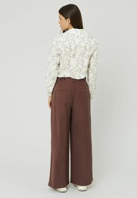 Blouse blanche à motifs floraux avec des manches longues, associée à un pantalon brun ample avec deux poches arrière et une fermeture à deux boutons.
