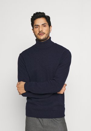 Marineblå turtleneck genser i strukturstrikk, med ribbestrikkede mansjetter og kant. Brukt med grå bukser, armene krysset. Enkelt design, ingen ekstra detaljer.