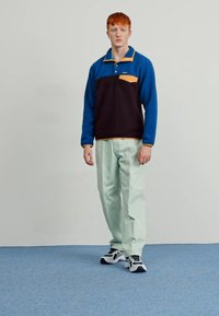 Fleece pullover in navy and blue with a contrasting orange pocket; paired with light green pants and black sneakers. Textured fabric.