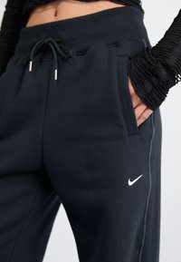 Mugavad mustad dressipüksid pehmest kangast, millel on nööriga vöökoht, küljetaskud ja väike valge Nike logo vasakul jalal.