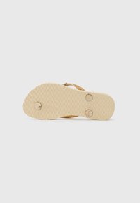 Beige leipäflip-flop, jossa on teksturoitu kumipohja. Koristeena kultainen kimalteellinen nauha ja kaksi pyöreää niittiä pohjassa lisäämään yksityiskohtia.