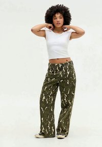 Calças largas em camuflagem verde com padrão abstrato bege, material de algodão, cintura alta e bolsos frontais. Combinadas com um top curto branco.