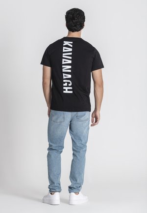 Black cotton t-shirt featuring vertical white text reading "KAVANAGH" on the back. Worn with light blue jeans and white sneakers.