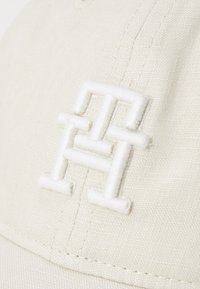 Vit basebollkeps i texturerat tyg med en broderad logotyp i vitt tråd. Sömmarna syns längs med sömmarna.