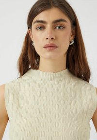 Top sin mangas de color beige con un patrón tejido texturizado. Presenta un escote alto y líneas horizontales sutiles para añadir detalle.