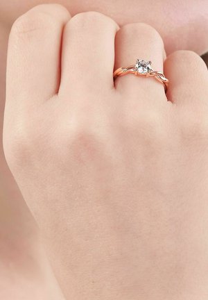 Hand wearing rose gold ring with a single round clear gemstone on the ring finger against a neutral background.