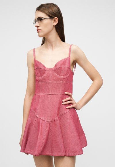 Femme portant une robe en denim rose sans manches avec un corsage structuré et une jupe évasée, regardant vers la gauche avec la main sur la hanche, sur un fond uni.
