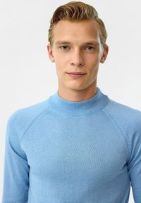 Lichtblauwe gebreide trui met een hoge hals, raglanmouwen en een gladde textuur. Kenmerkt zich door een nauwsluitende pasvorm en subtiele stikseldetails.