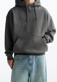 Sudadera gris oscuro con bolsillo tipo canguro, cordón ajustable y mangas largas. Combinada con pantalones de mezclilla de color azul claro. Textura suave, corte relajado.