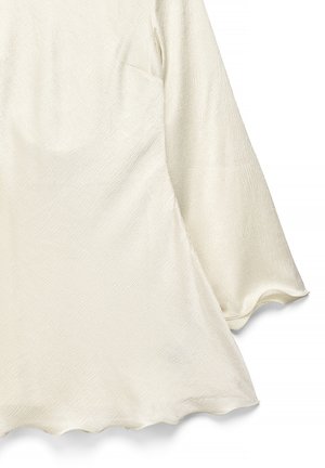Top color crema hecho de tela texturizada, con mangas acampanadas largas y dobladillo en forma de conchas. La superficie tiene un patrón sutil.