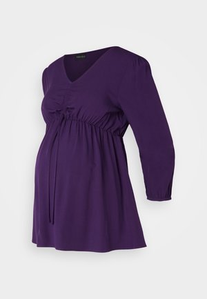 Fioletowa bluzka ciążowa z dekoltem w serek, długimi bufiastymi rękawami, marszczonym materiałem w pasie i luźnym krojem zapewniającym komfort i swobodę podczas ciąży.