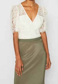 Vit spetsblus med puffärmar, djup V-hals och olivgrön satinkjol; elegant design med blommönster och slät textur.