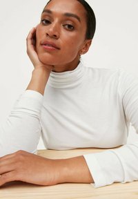 Hvid turtleneck top i blødt stof med lange ærmer. Model hviler hagen på hånden og viser en glat, neutral-tonet hudtekstur.