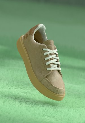 Tan suede sneaker with white laces, rounded toe, and a light tan rubber sole. Features a logo on the heel and a soft interior lining.