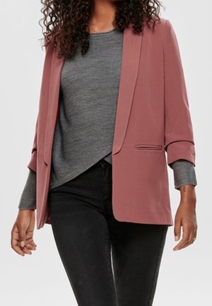 Femme portant un blazer rose poudré sur un haut côtelé gris et un pantalon noir, debout, les bras détendus le long du corps.